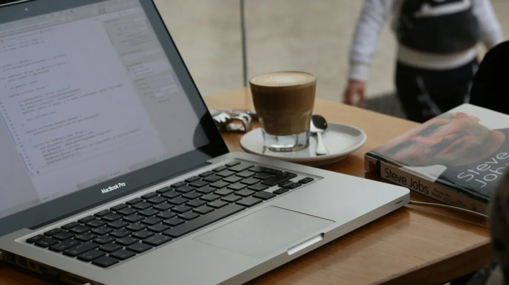 MacBook Pro s otvoreným Xcode kódom, káva a kniha Steve Jobs na stole